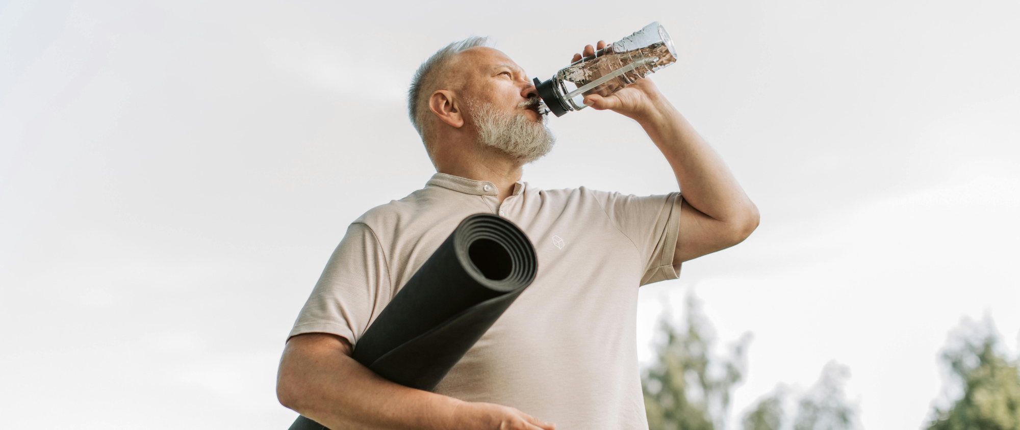 Älterer Mann mit Yogamatte trinkt Wasser aus einer Flasche im Freien.