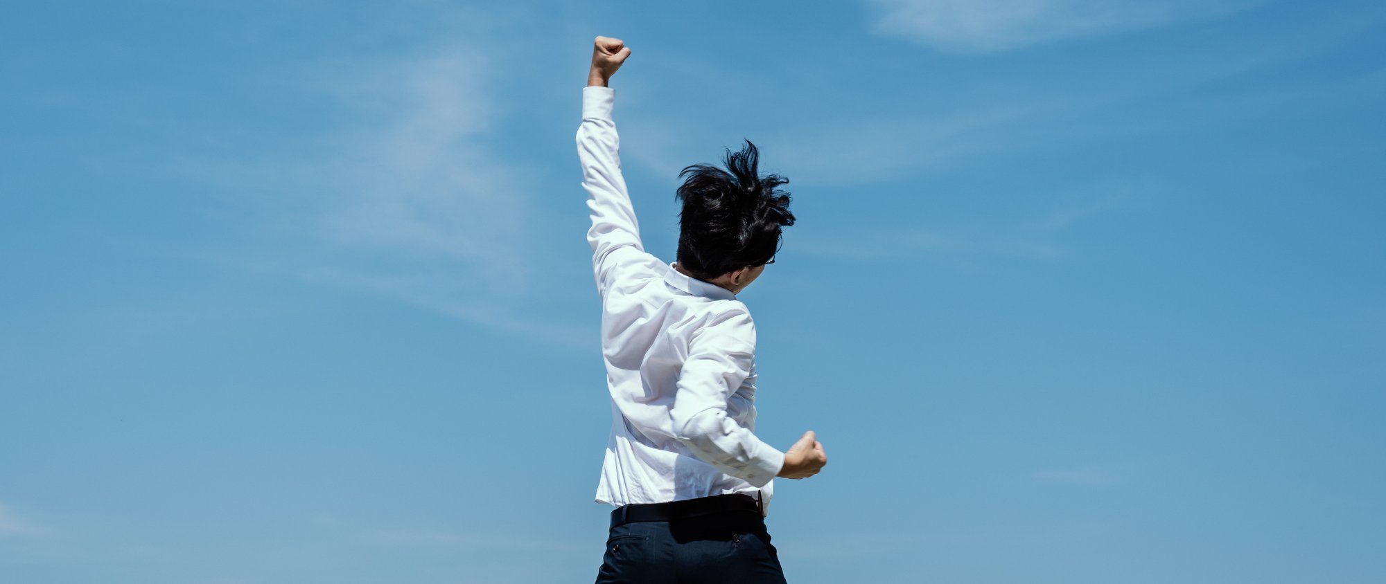 Mann streckt jubelnd die Faust in die Luft vor blauem Himmel.