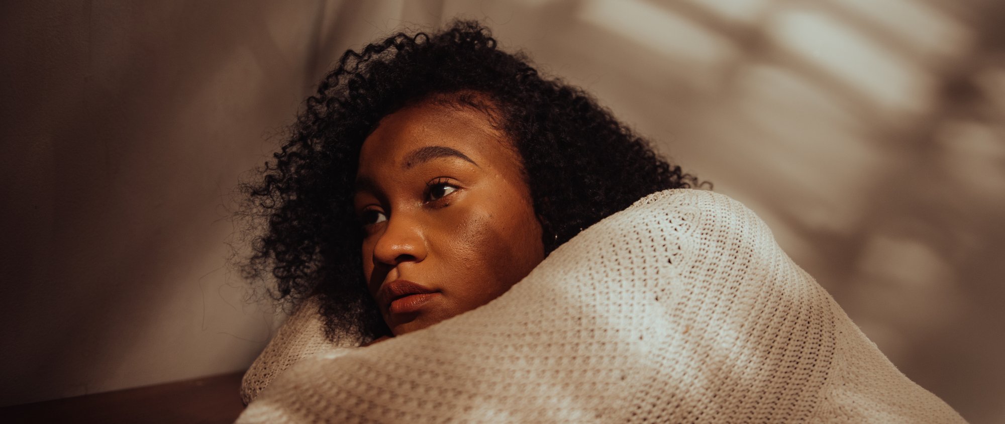 Nachdenkliche Frau eingehüllt in eine Decke sitzt im Halbschatten.