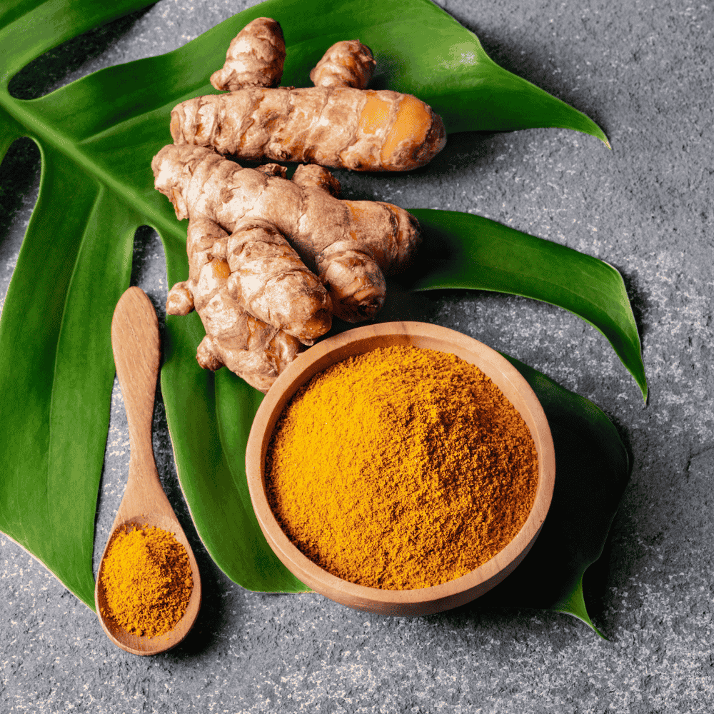 Kurkuma-Wurzel mit daneben liegendem orangefarbenem Kurkumapulver in einer Holzschale und auf einem Löffel. Die Elemente liegen auf einem grünen Blatt